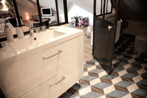 Une salle de bain moderne avec un meuble-lavabo blanc, un évier et un miroir. Le sol présente un motif à carreaux et on peut apercevoir un petit chien à l’arrière-plan.