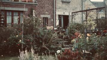 Un jardin avec différentes plantes et fleurs, un bâtiment en briques et un portail en métal.