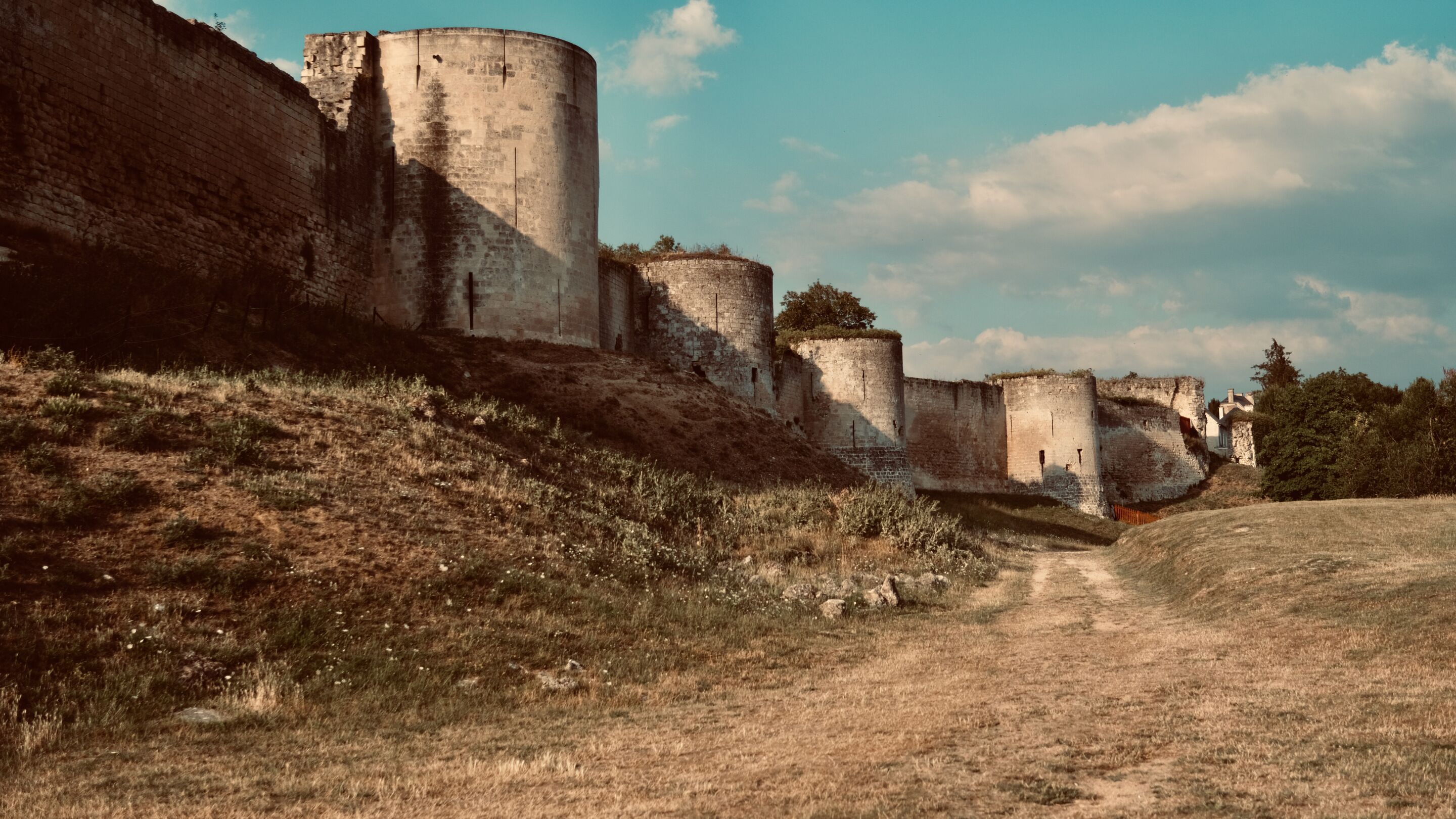 A historical stone structure with multiple cylindrical towers, a grassy foreground, and a clear sky.