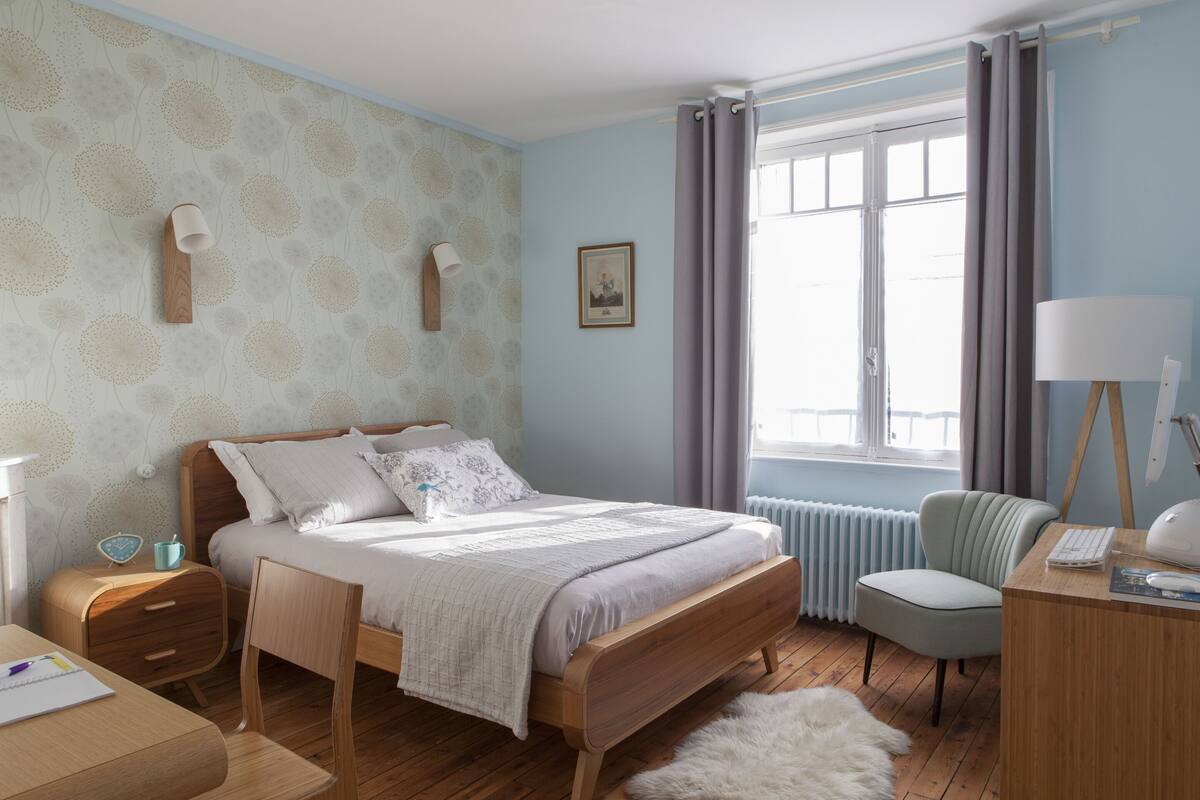 A bedroom with a wooden bed, a desk with a lamp, a chair, a radiator, and a window with curtains.