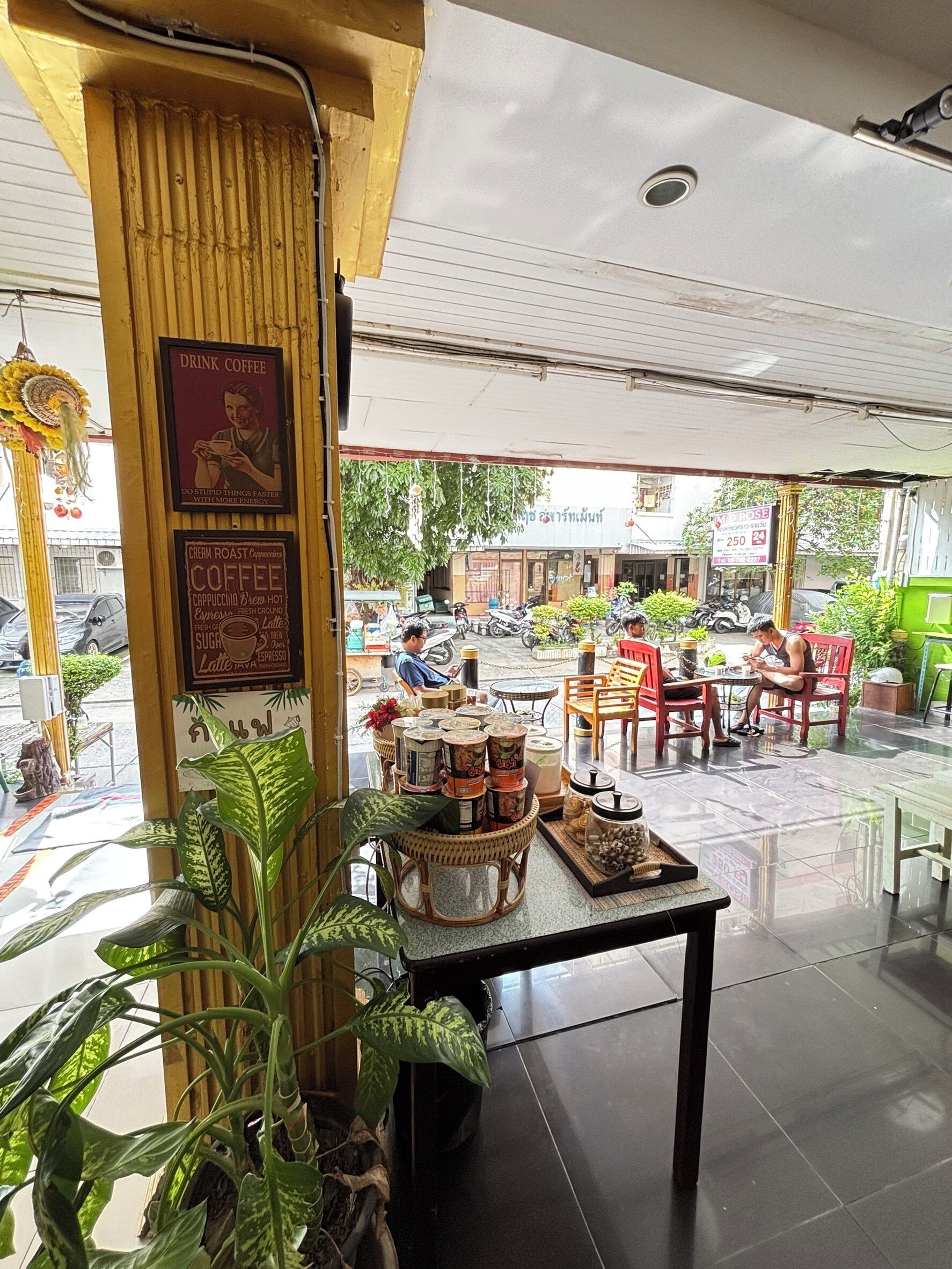 Un café al aire libre con mesas y sillas, diversas plantas y suelo de baldosas.