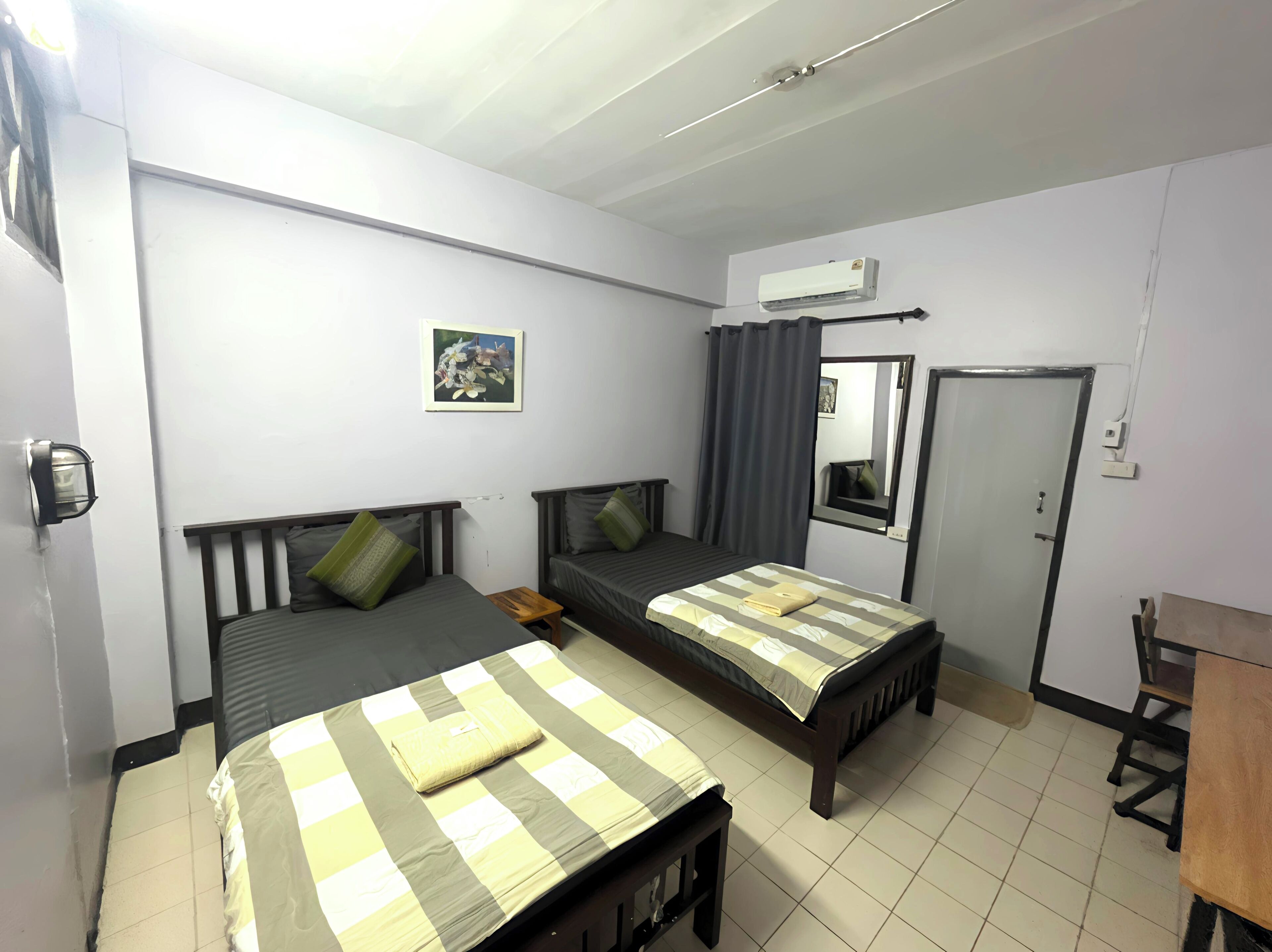 A room with two beds, a small table, an air conditioning unit, and a mirror on the wall.