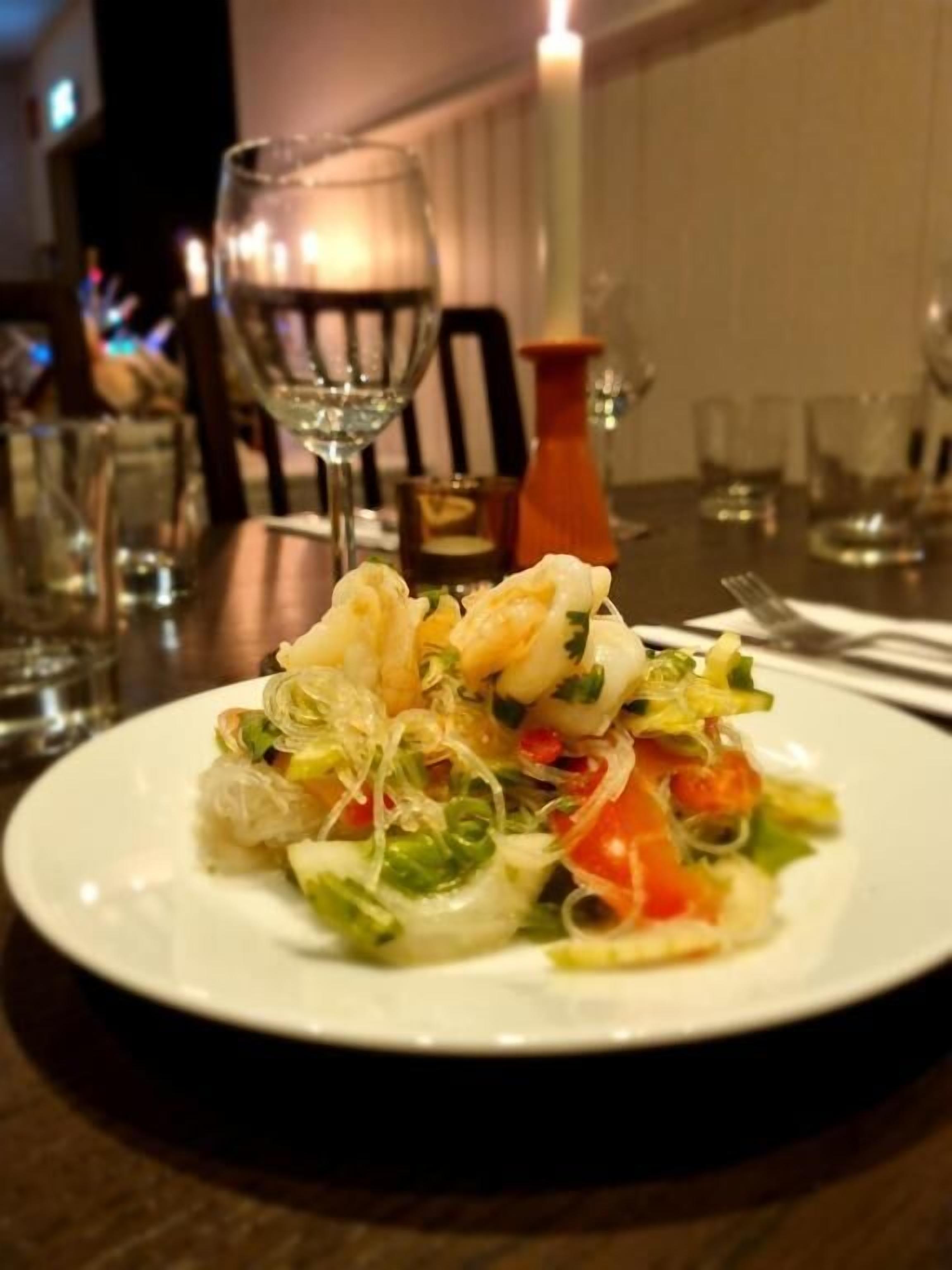 A plate of food with shrimp and vegetables on a white plate, set on a table with glasses and a candle.