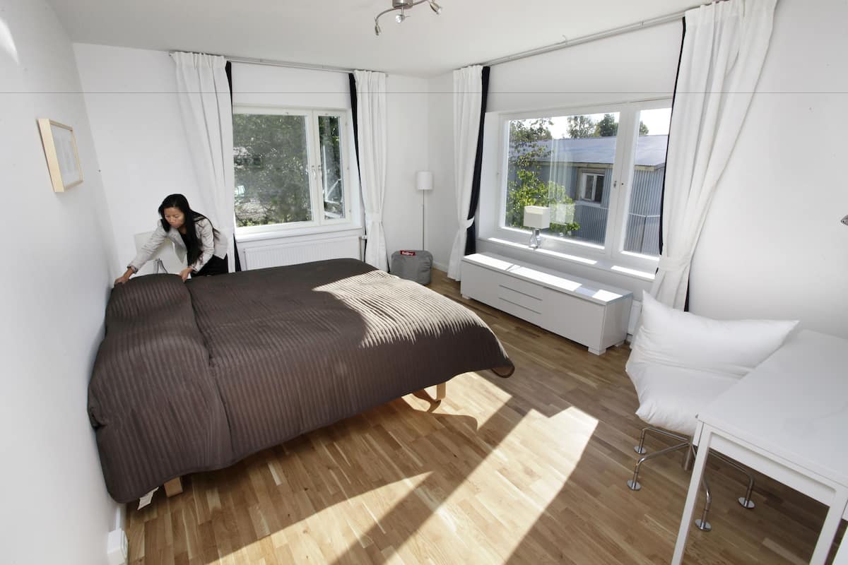 A bedroom with a large bed, a bedside table, a window with curtains, and a view of the outside.