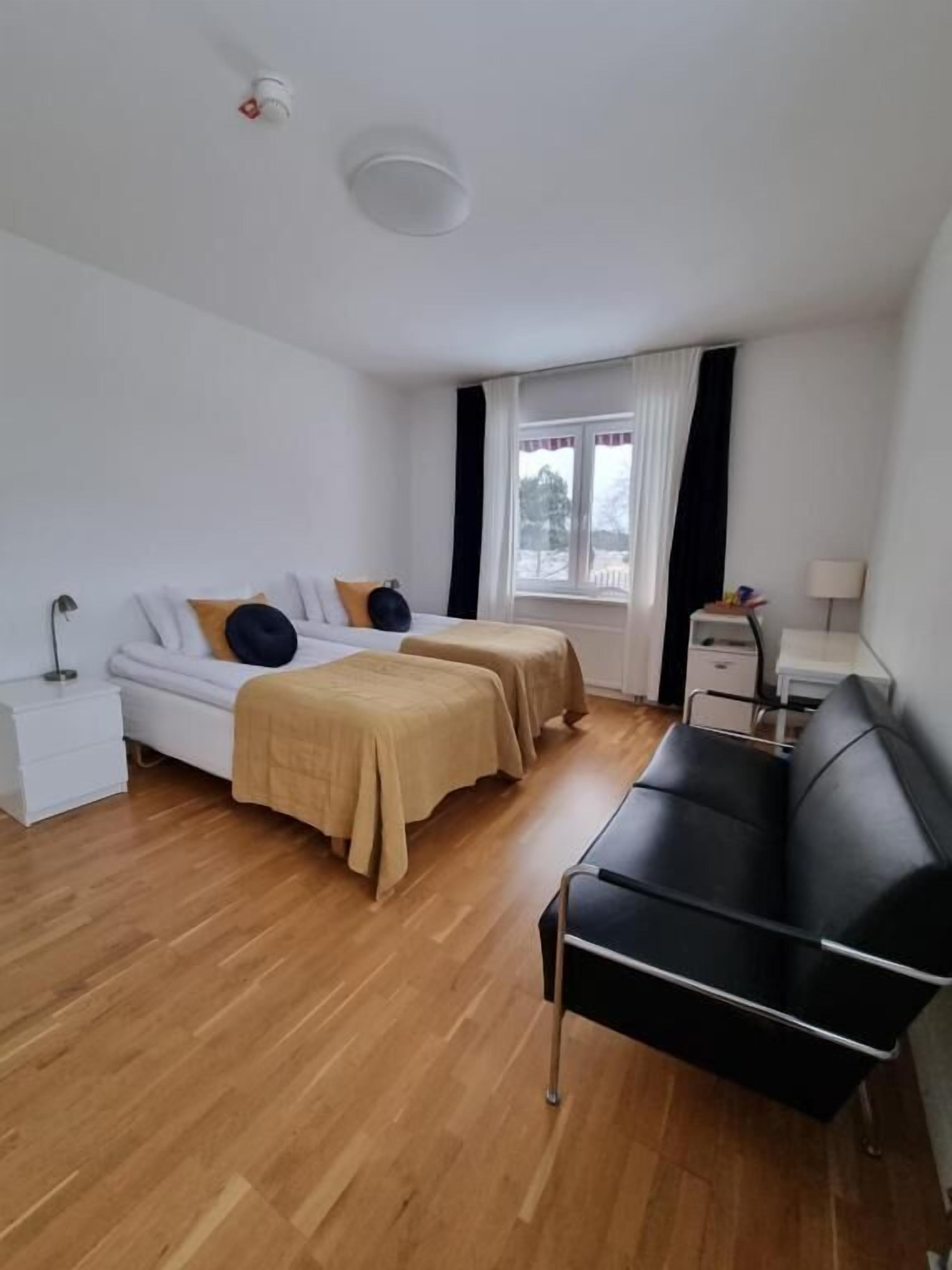 Ett modernt hotellrum med trägolv, två sängar med beige och svart sängkläder, en svart soffa, ett skrivbord och ett fönster med gardiner.