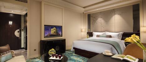 A hotel room with a large bed, a desk with a chair, a small table with a vase of flowers, and a television on a stand.