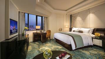 A hotel room with a large bed, a desk with a laptop, a TV on the wall, and a view of the city at dusk.