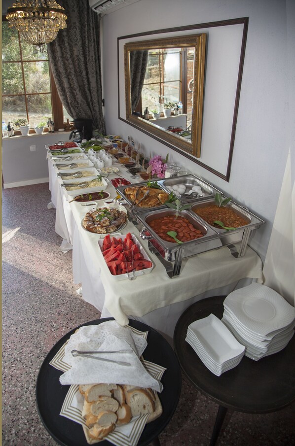A buffet table with various food items and utensils, a mirror, and a window with curtains. - Iola Hotel (Bozcaada)