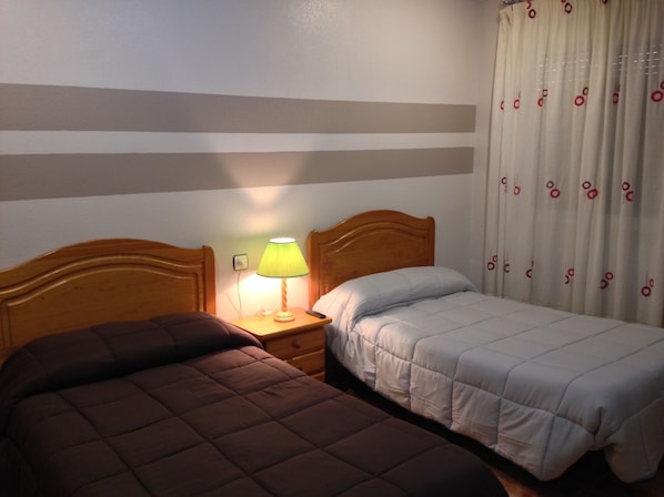 Habitación de hotel con dos camas, una mesita de noche con lámpara y una cortina estampada.