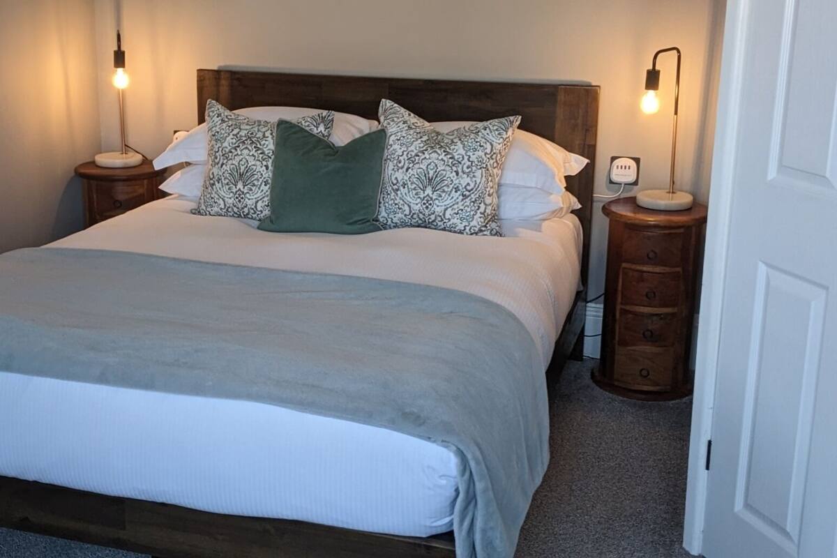 A neatly made bed with a grey blanket, a wooden headboard, and patterned pillows. There are two bedside tables with lamps, one on each side of the bed.