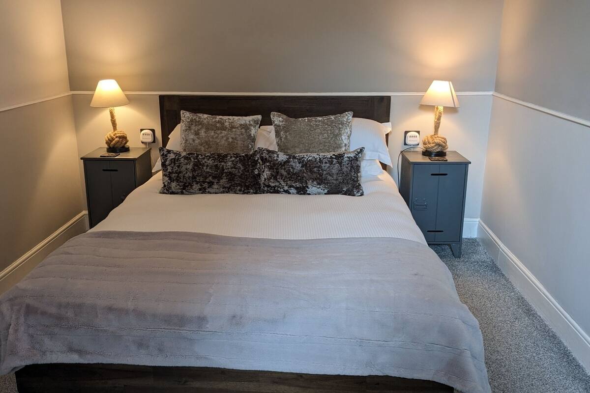 A neatly made bed with a grey duvet, two bedside tables with lamps, and a dark headboard.