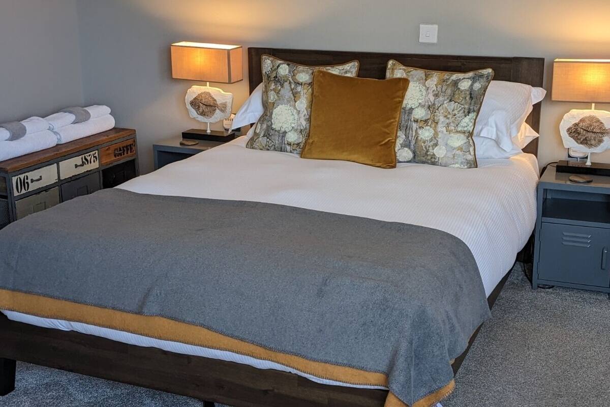 A neatly made bed with a grey and brown blanket, two bedside lamps, a wooden nightstand with a lamp, and a small cabinet.