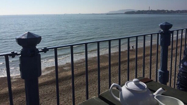 Blick auf das Meer vom Balkon mit Geländer, ein Tisch mit Teekanne und Tassen sowie eine entfernte Küstenlinie.