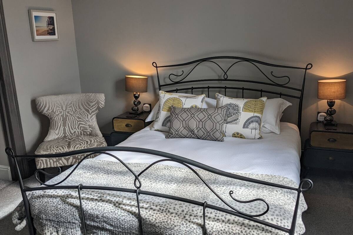 A neatly made bed with a metal frame, two bedside tables with lamps, a chair, and a framed picture on the wall.