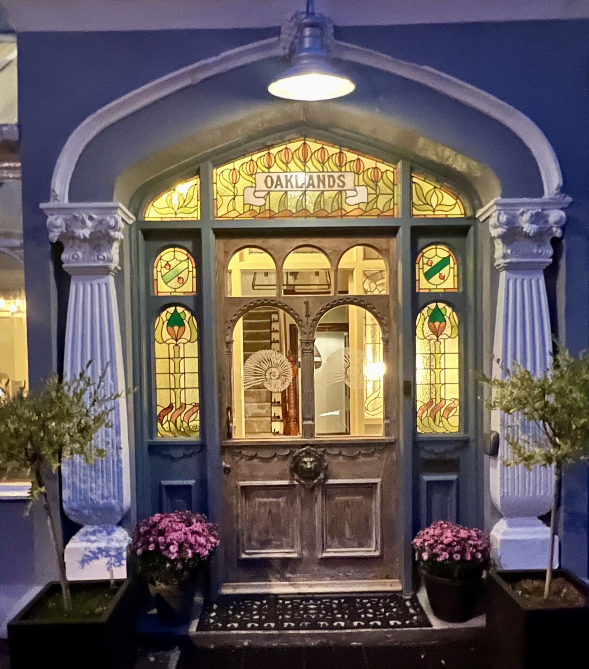 L’entrée d’un immeuble, avec une enseigne portant l’inscription « OAKLANDS » au-dessus de la porte, flanquée de deux colonnes et agrémentée de plantes en pot.