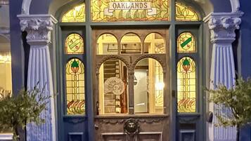 L’entrée d’un immeuble, avec une enseigne portant l’inscription « OAKLANDS » au-dessus de la porte, flanquée de deux colonnes et agrémentée de plantes en pot.