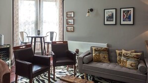 Ein Wohnzimmer mit einer Couch, zwei Sesseln, einem Couchtisch und einer Chandelier. Drei gerahmte Bilder hängen an der Wand und es gibt ein Fenster mit Vorhängen.