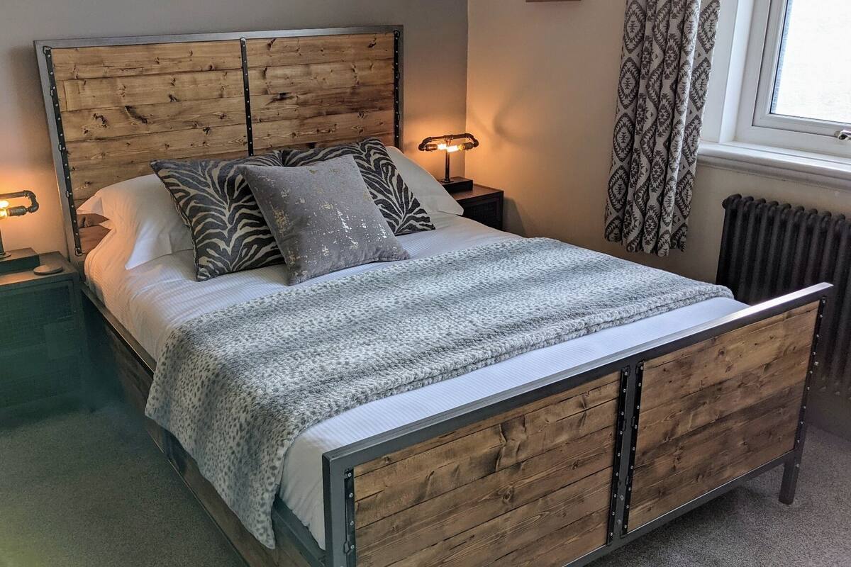 A bedroom with a wooden headboard, a bed with patterned pillows, a grey blanket, bedside lamps, and a window with patterned curtains.