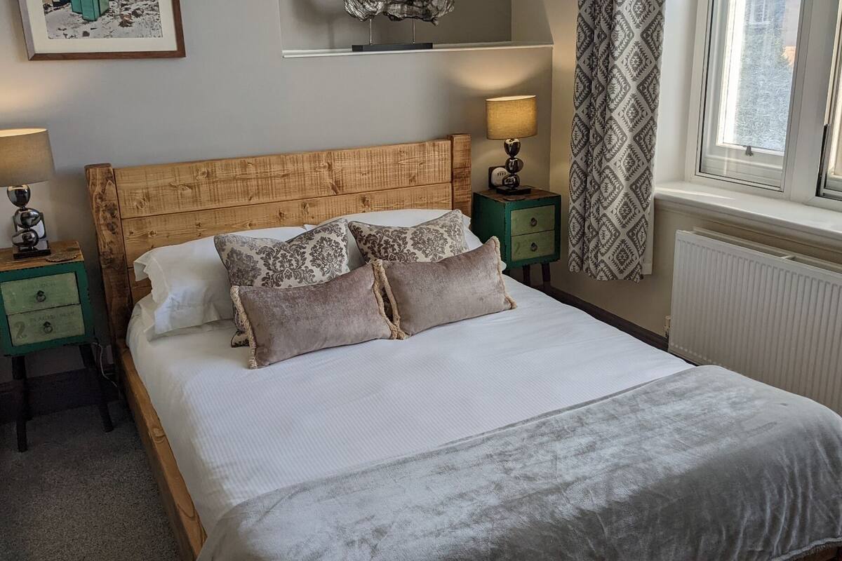 A bedroom with a wooden headboard, two bedside tables, a bed with white bedding, patterned pillows, a window with curtains, and a radiator.