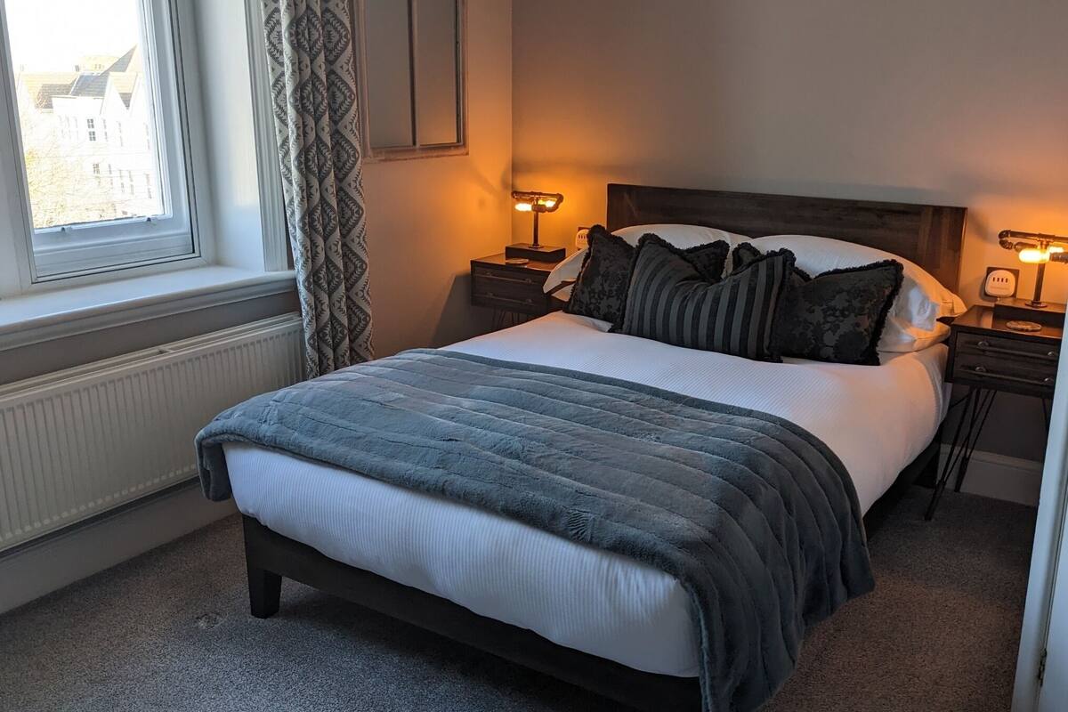 A neatly made bed with a dark headboard, two bedside lamps, and a window with curtains.