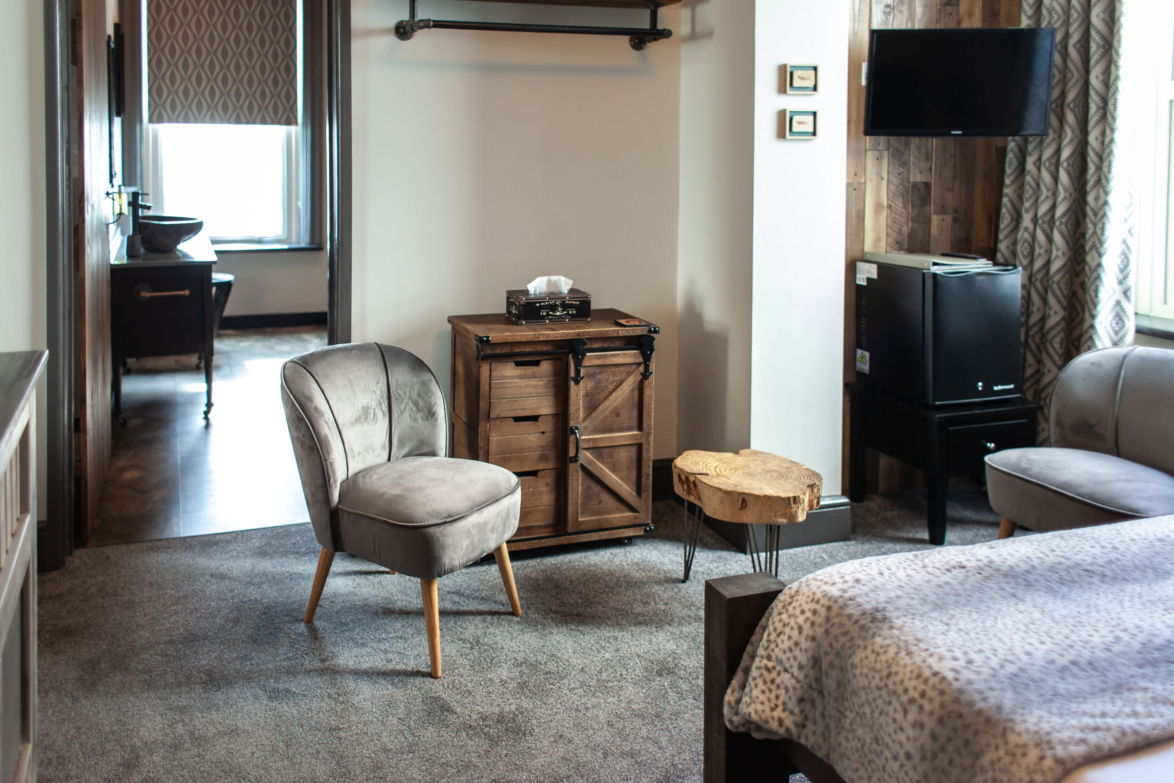 A hotel room with a grey armchair, a wooden chest of drawers, a small round wooden table, a television, and a bed with a patterned bedspread.