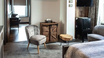 A hotel room with a grey armchair, a wooden chest of drawers, a small round wooden table, a television, and a bed with a patterned bedspread.