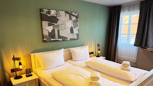 A double bed with white linens and two rolled towels, a bedside table with a lamp, a nightstand, and a wall-mounted abstract painting. - Apartments Casa da Enzo (Goslar)