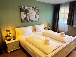Una cama doble con ropa blanca y dos toallas enrolladas, una mesita de noche con lámpara, una mesita auxiliar y un cuadro abstracto colgado en la pared.