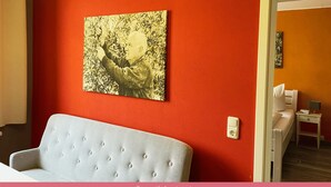 A room with a red wall, a framed artwork, a light grey sofa, and a bedside table with a lamp. - Apartments Casa da Enzo (Goslar)