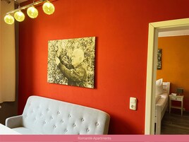 Una habitación con una pared roja, una obra de arte enmarcada, un sofá gris claro y una mesita de noche con una lámpara.