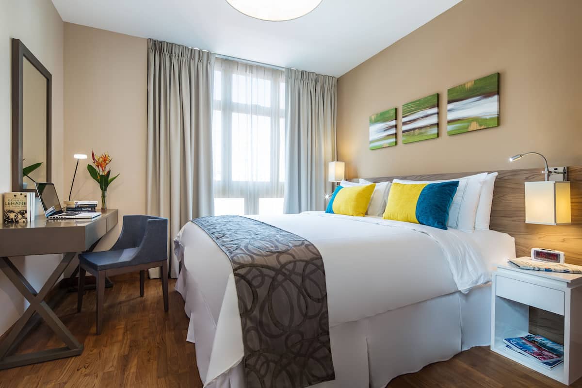 A hotel room with a bed, desk, chair, lamp, and artwork on the wall.