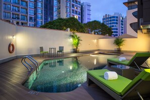 Una piscina en la azotea con sillones de descanso y un jacuzzi, rodeada de edificios modernos y árboles.