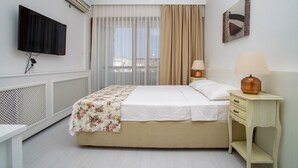 Quarto de hotel com cama, televisão fixada na parede, mesa de cabeceira com abajur e janela com cortinas.