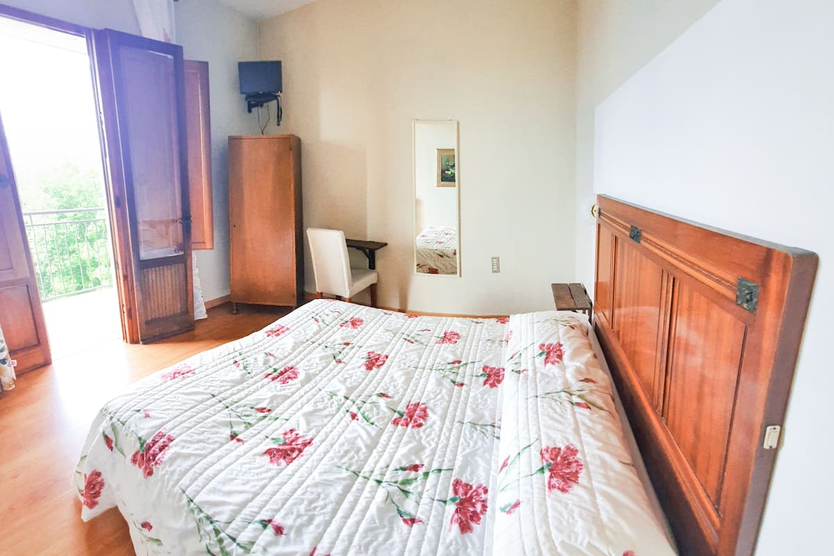 A bedroom with a wooden headboard, a bed with a floral quilt, a wooden wardrobe, a chair, and a television on a stand.