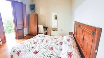 Un dormitorio con cabecera de madera, cama con colcha floral, armario de madera, una silla y un televisor sobre un mueble bajo.