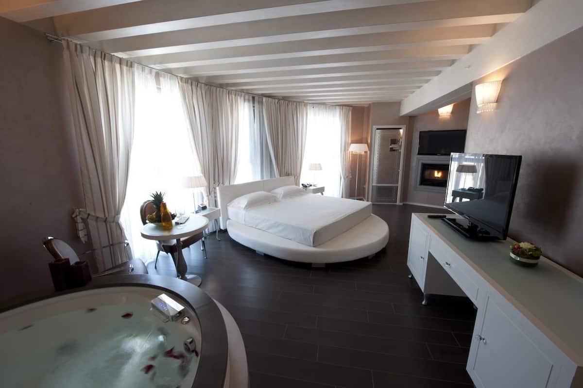 A modern hotel room with a large bed, a TV, a fireplace, a bathroom with a bathtub, and a dining area.