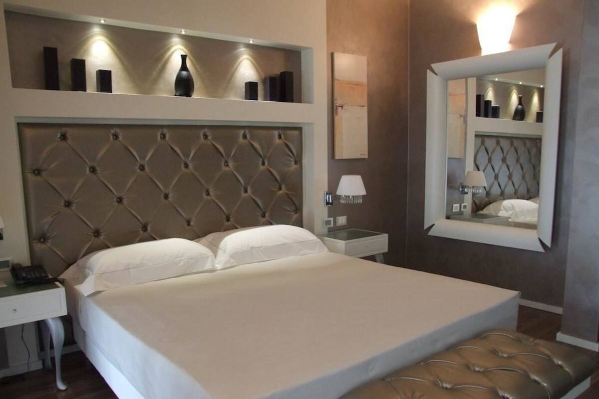 A neatly arranged hotel room with a large bed, a bedside table, a nightstand, a mirror, and a small shelf with decorative items.