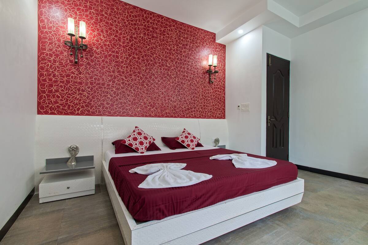 A modern hotel room with a large bed, red patterned wall, two wall sconces, and a dark door.