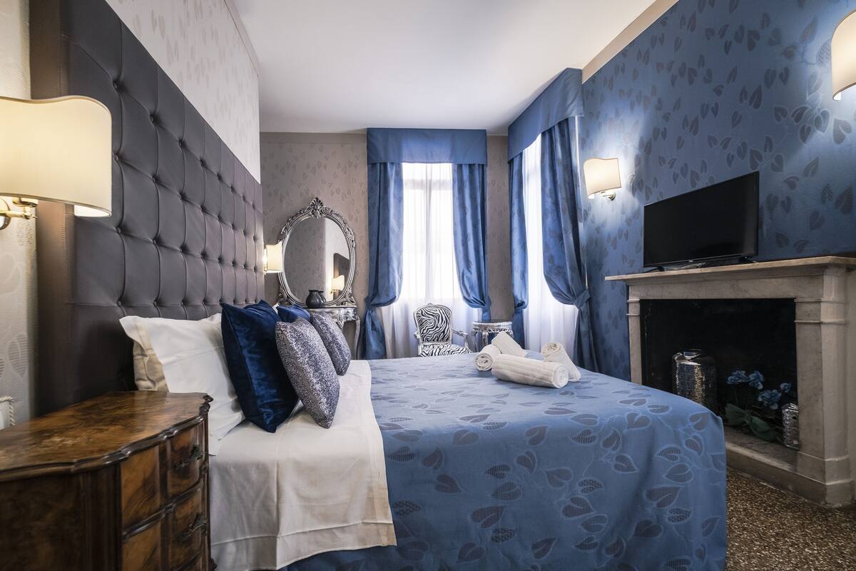 A hotel room with a tufted headboard, a large mirror, a flat-screen TV, a fireplace, and a bed with a blue patterned bedspread.