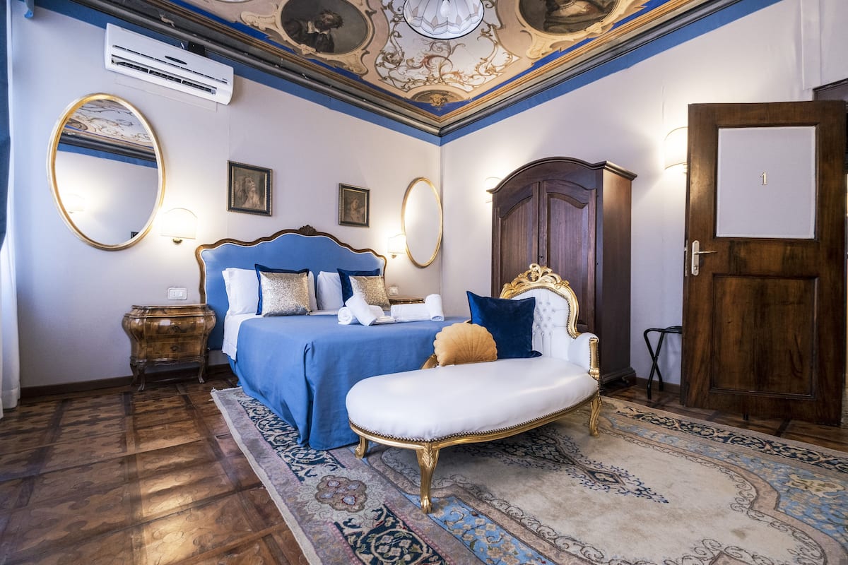 A room with a large bed, a sitting chair, a wardrobe, and a mirror.