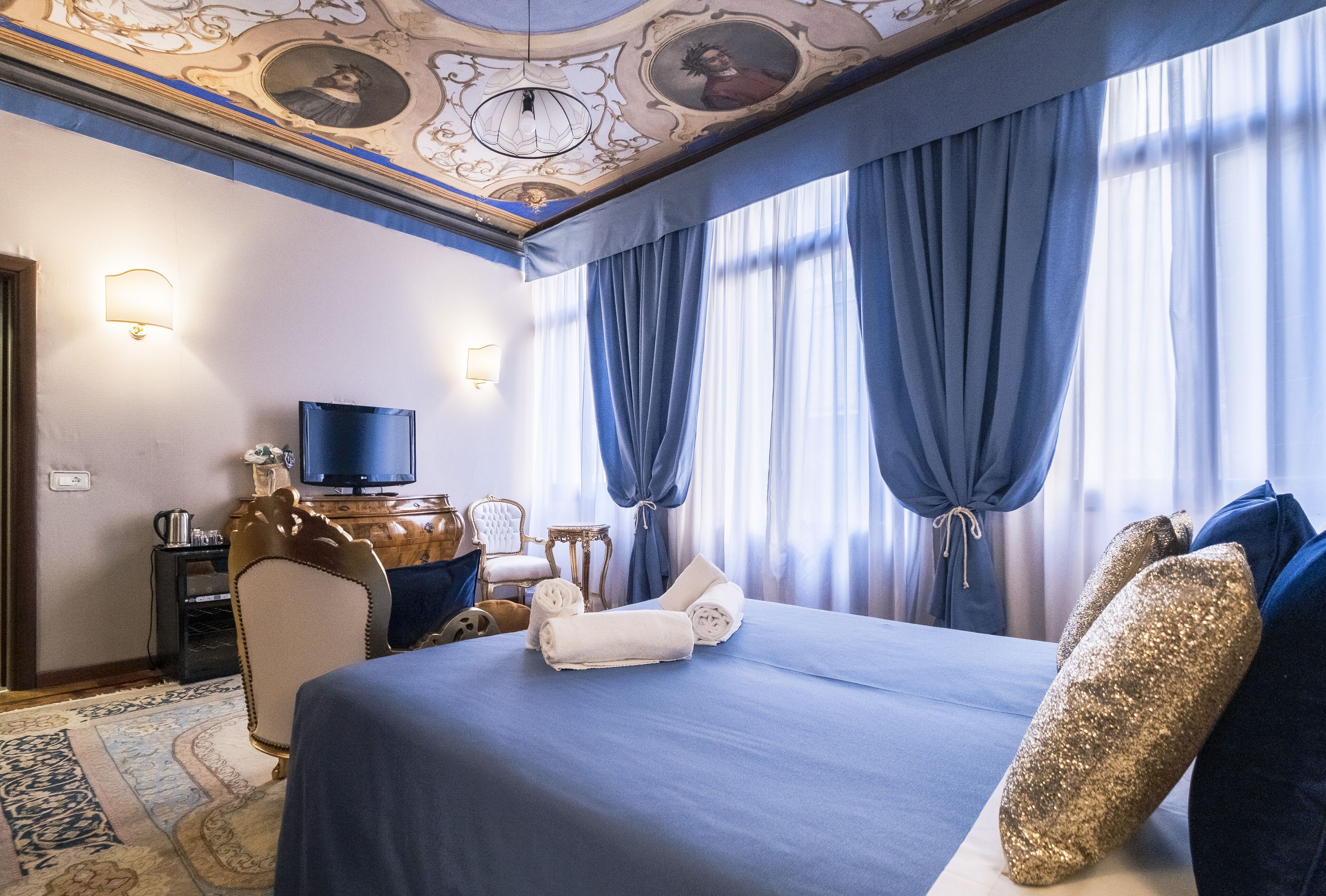 A hotel room with a large bed, a television, a desk with a lamp, and a decorative ceiling.