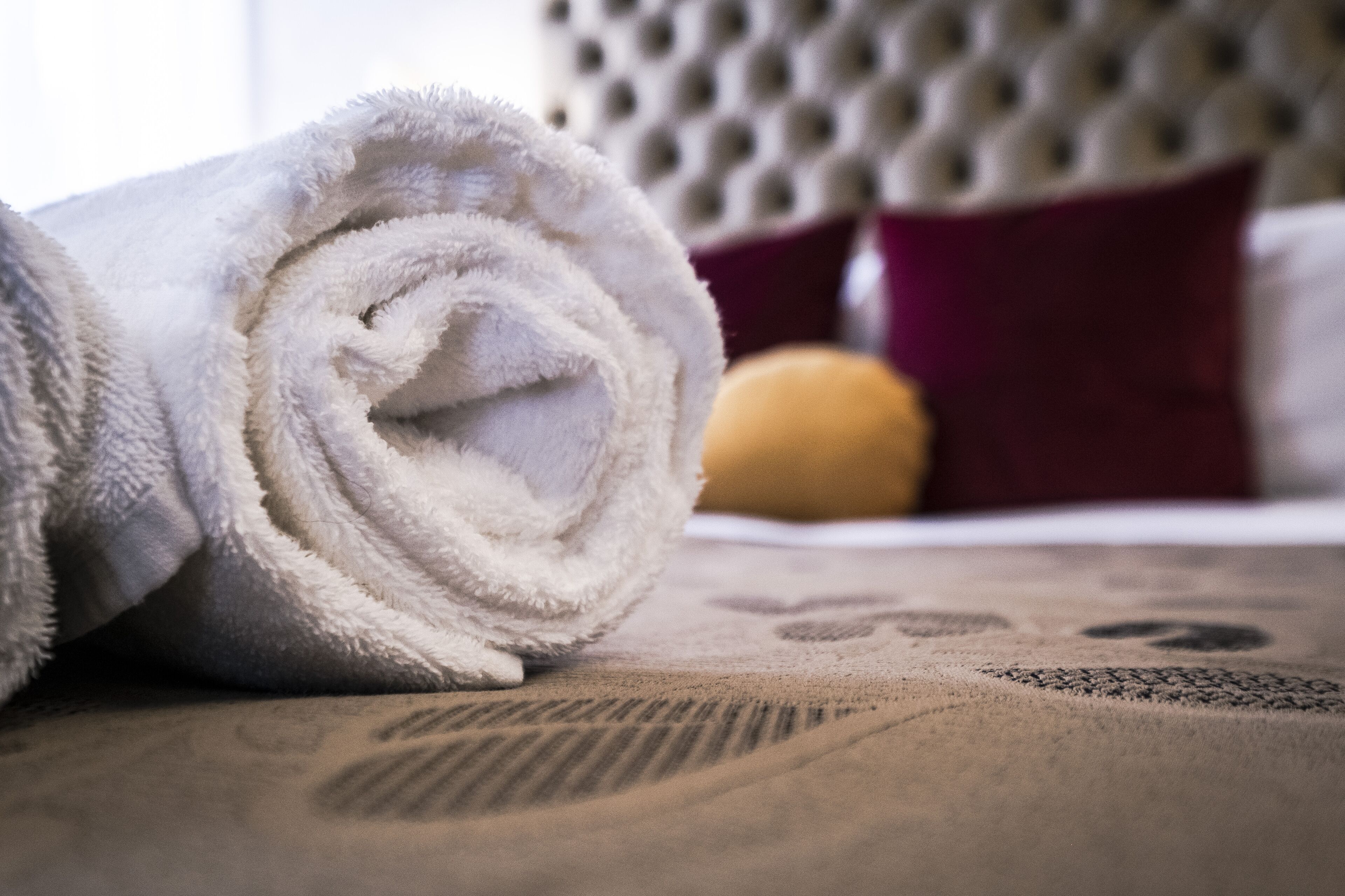 Een opgerolde witte handdoek op een bed met een getextureerd sprei en een gebordureerd hoofdeinde, met een rode kussen op de achtergrond.
