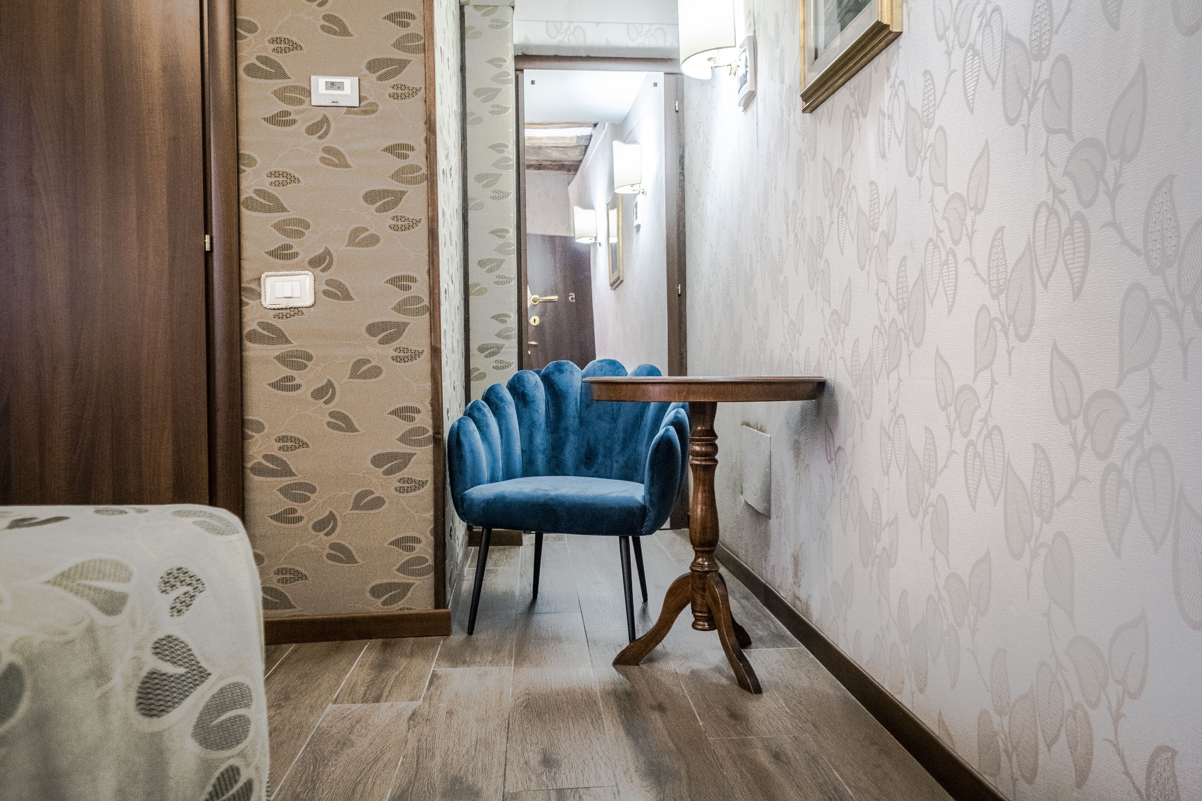 A room with a wooden wardrobe, a blue chair, a wooden table, and patterned wallpaper.