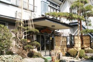 Un bâtiment de style japonais traditionnel, doté d’une entrée couverte, entouré de bonsaïs bien entretenus et d’un sentier en pierre.