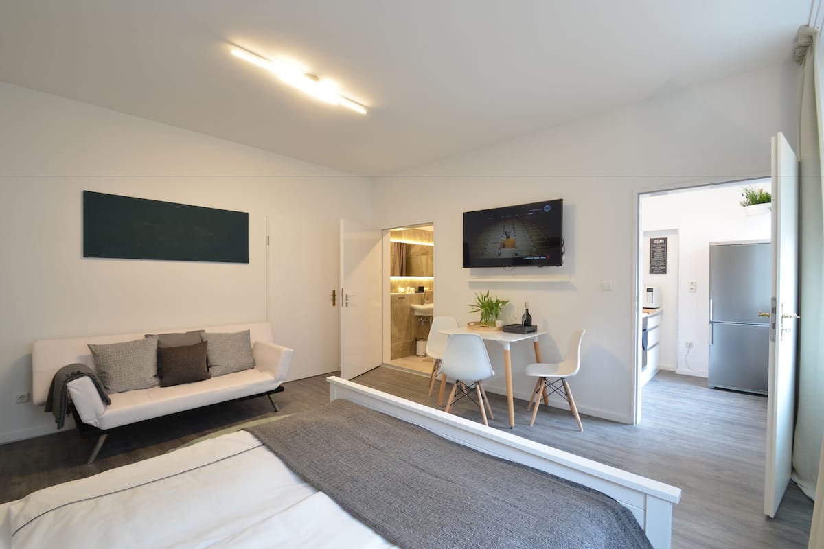 Habitación moderna y luminosa con cama, sofá, mesa de comedor con sillas, televisor montado en la pared y una cocina visible a través de una puerta abierta.