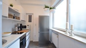 Cocina moderna con armarios blancos, un refrigerador de acero inoxidable, un microondas y una cafetera. Hay plantas en macetas en el alféizar de la ventana y una ventana con vistas.