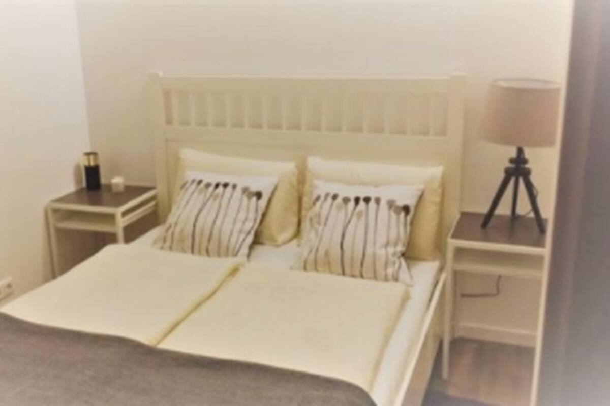 Un dormitorio con una cama, dos mesitas de noche, una lámpara y una pequeña mesa con un trípode.