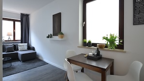 Una sala moderna con un sofá, una mesa de comedor con sillas, una ventana con plantas y un estante montado en la pared con objetos decorativos.