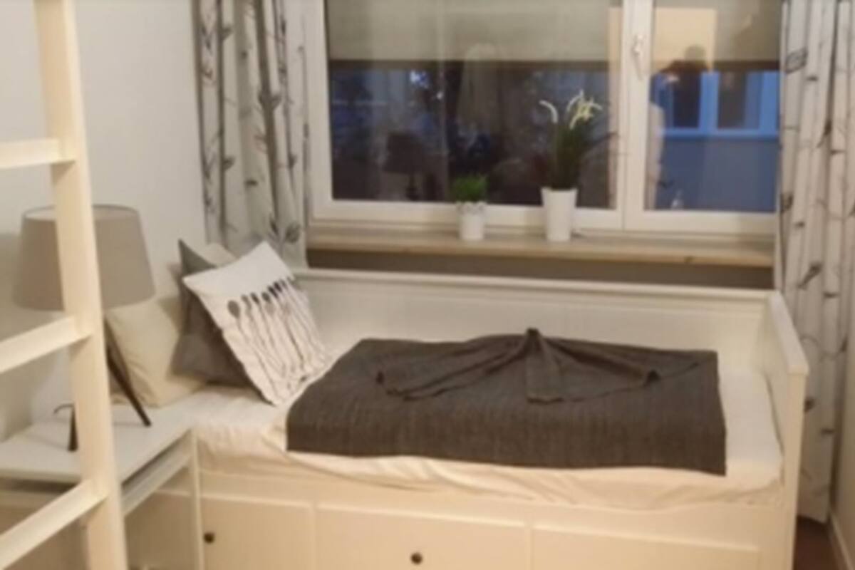 Una habitación pequeña y acogedora con una cama individual, una ventana con vistas al exterior y una estantería con una lámpara.