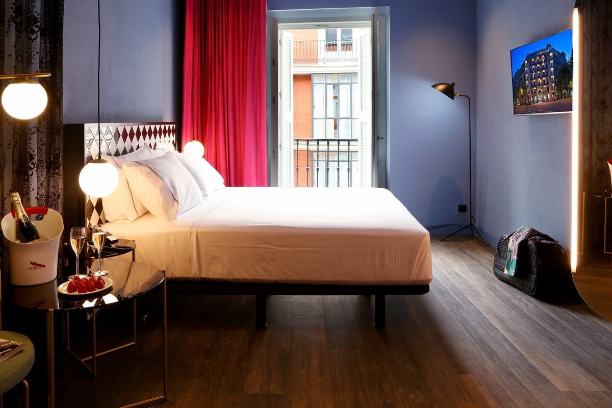 Habitación de hotel con una cama, una mesita de noche con una botella de champán, una copa de champán y fresas. Hay una silla con una revista, una lámpara y un cuadro en la pared.